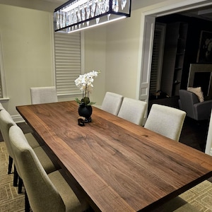 Peut inclure: Une salle à manger avec une grande table rectangulaire en bois foncé, entourée de chaises rembourrées. Au-dessus de la table, un lustre rectangulaire avec des cristaux. Un vase de fleurs blanches est posé sur la table.