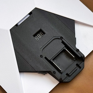 Peut inclure: Un appareil électronique noir avec un affichage numérique indiquant "8.8.8" est partiellement visible, encadré par du papier blanc. L'appareil a une surface texturée et une ouverture rectangulaire avec des contacts métalliques. Le fond est un matériau brun semblable à du cuir.