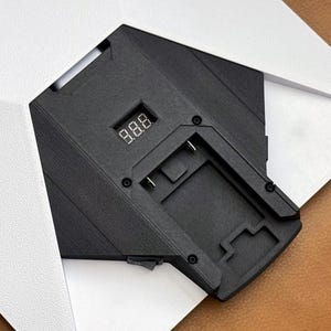 May include: A black electronic device with a digital display showing "8.8.8" is partially visible, framed by white paper. The device has a rectangular shape with various geometric features and small circular details.