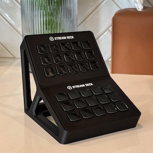 May include: Black Stream Deck with multiple square buttons. The device has the Stream Deck logo and text printed on it. The device is angled and sits on a light-colored surface.