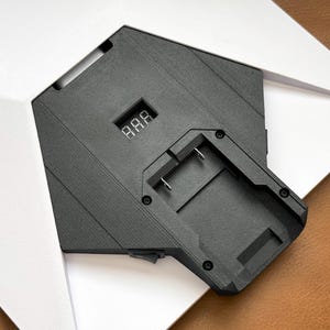 Peut inclure: Une station de charge de batterie noire avec un écran d'affichage numérique montrant trois chiffres, montée sur une base géométrique blanche. Le chargeur dispose d'une fente encastrée avec deux broches de contact en métal et un interrupteur noir.