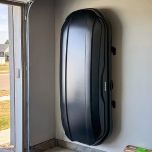 May include: A black Yakima cargo box mounted on a garage wall. The box is oblong and has a smooth, reflective surface. The Yakima logo is visible on the side. The box is secured with black mounting brackets.