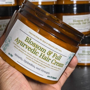 May include: A jar of Blossom & Full Ayurvedic Hair Cream, 12 oz, with a white label featuring text and floral accents. The label lists ingredients and highlights growth, hydration, and strength. The jar is held in a hand, with other jars in the background.