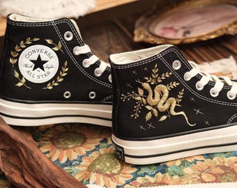 May include: A pair of black high-top Converse sneakers with gold embroidered details. One shoe features a snake and floral design, while the other has a classic Converse All Star logo with a laurel wreath.