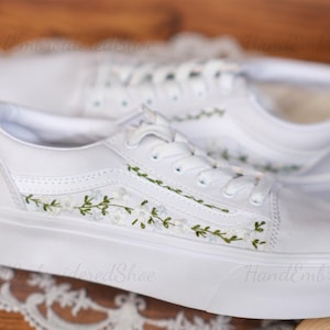 Könnte beinhalten: Weiße Canvas-Sneaker mit handgefertigten grünen Blumenstickereien. Die Schuhe haben eine weiße Gummisohle und sind im klassischen Stil gehalten.