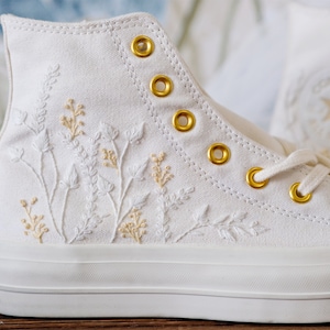 May include: White high-top canvas sneakers with gold eyelets and embroidered floral details in white and cream.