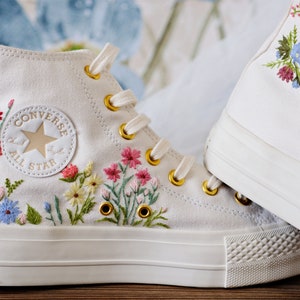 Puede incluir: Zapatillas Converse blancas de caña alta con flores bordadas en rosa, azul y amarillo. El logotipo de Converse All Star es visible en el lateral de la zapatilla.