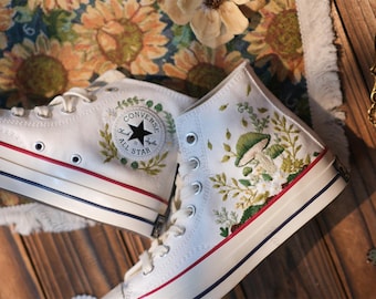 May include: White high-top Converse sneakers with embroidered floral designs, including mushrooms and leaves. The Converse logo is visible on one shoe.