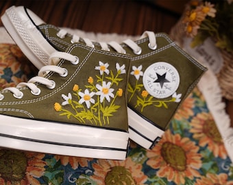 May include: Olive green high-top sneakers with white laces and soles. The sneakers feature embroidered white daisies with yellow centers. A Converse All Star patch is on the side.
