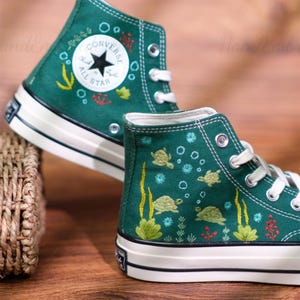 May include: Green high-top sneakers with white laces and soles. The sneakers feature embroidered sea turtles, bubbles, and aquatic plants. The Converse All Star logo is visible on the side.