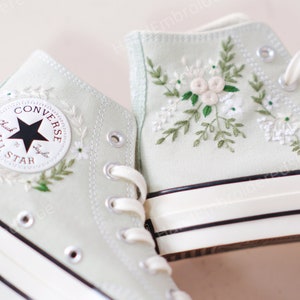 Puede incluir: Un par de zapatillas Converse de caña alta de color verde claro con cordones blancos y un logotipo de estrella negra. Las zapatillas están bordadas con flores blancas y hojas verdes.