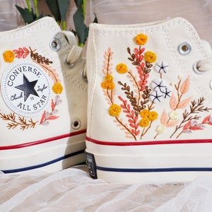 May include: Cream-colored high-top sneakers with floral embroidery. One shoe features a Converse All Star logo with a floral wreath. The other shoe has a design of yellow, orange, and brown embroidered flowers and leaves. The soles have red and blue stripes.
