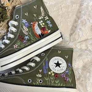 May include: A pair of green canvas high-top sneakers with embroidered flowers, a fox, and bees. The sneakers have white soles and a black Converse All Star logo.