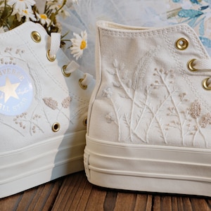May include: White high-top Converse sneakers with embroidered floral designs and gold eyelets. The sneakers have a platform sole and the Converse All Star logo is visible on the side.