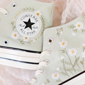 Puede incluir: Un par de zapatillas Converse de caña alta de color verde claro con flores blancas bordadas y una estrella negra en el lateral. El texto "Converse Chuck Taylor All Star" está en el lateral de la zapatilla.