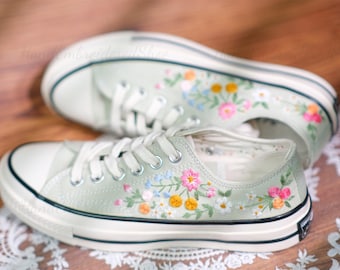 May include: A pair of light green canvas sneakers with white laces and toe caps. The sneakers are adorned with colorful embroidered flowers in shades of pink, yellow, and blue. The text "Handembroidered Shoe" is visible on the top of the shoe.