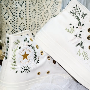 May include: White high-top Converse sneakers with green floral embroidery and a gold star logo. The text "Converse All Star" is embroidered on the side of the shoe.