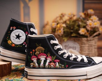 May include: Black high-top Converse sneakers with white stitching and a white rubber sole. The sneakers are embroidered with a floral design and mushrooms.