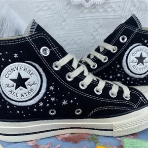 May include: Black high-top Converse sneakers with white stitching and a crescent moon and stars design. The Converse logo is embroidered on the side of the shoe.