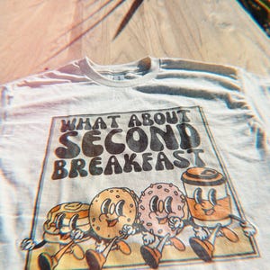 Unisex beige shirt with a retro graphic of breakfast foods and “What About Second Breakfast” text, perfect for LOTR fans.
