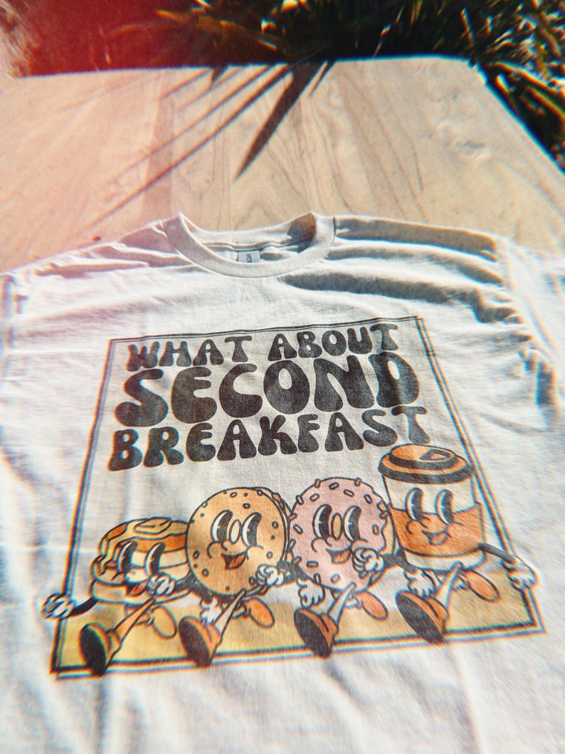 Unisex beige shirt with a retro graphic of breakfast foods and “What About Second Breakfast” text, perfect for LOTR fans.
