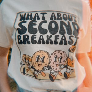 Beige retro t-shirt with vintage-inspired graphic of a donut, bagel, pancakes, and coffee, featuring the text “What About Second Breakfast.”