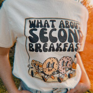 Beige retro t-shirt with vintage-inspired graphic of a donut, bagel, pancakes, and coffee, featuring the text “What About Second Breakfast” in a retro font. referring to the famous hobbit quote in the lord of the rings