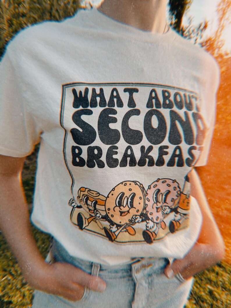 Beige retro t-shirt with vintage-inspired graphic of a donut, bagel, pancakes, and coffee, featuring the text “What About Second Breakfast” in a retro font. referring to the famous hobbit quote in the lord of the rings