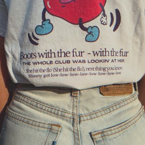 close up of the part of the back of the white t-shirt featuring a retro-style apple with boots and the full Flo Rida “Low” lyrics.