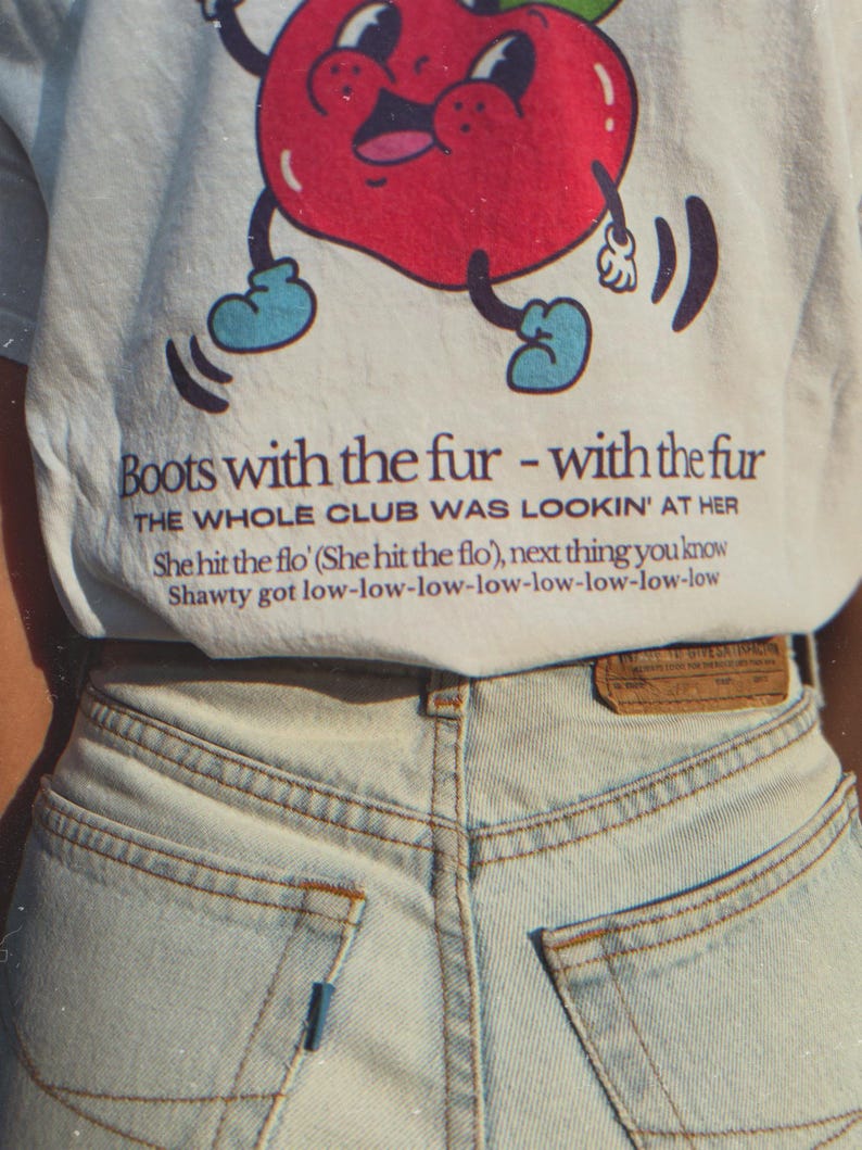 close up of the part of the back of the white t-shirt featuring a retro-style apple with boots and the full Flo Rida “Low” lyrics.