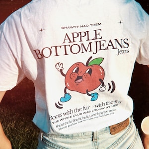 May include: White t-shirt featuring a red apple graphic and the text "SHAWTY HAD THEM APPLE BOTTOM JEANS Jeans" and "Boots with the fur - with the fur THE WHOLE CLUB WAS LOOKIN' AT HER She hit the flo (She hit the flo), next thing you know Shawty got low-low-low-low-low-low-low-low"