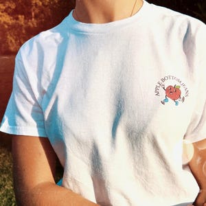 Front of white t-shirt with a small apple graphic on the chest and the text “Apple Bottom Jeans”, retro vibe design ideal for Flo Rida fans