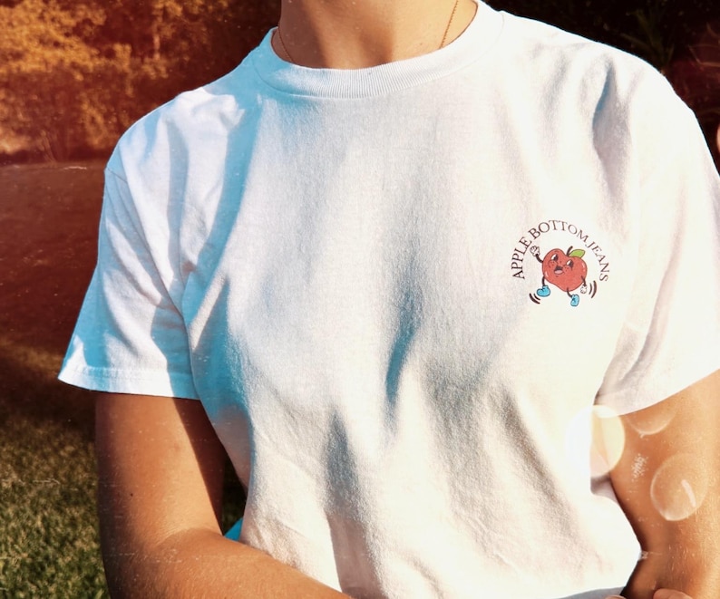 Front of white t-shirt with a small apple graphic on the chest and the text “Apple Bottom Jeans”, retro vibe design ideal for Flo Rida fans