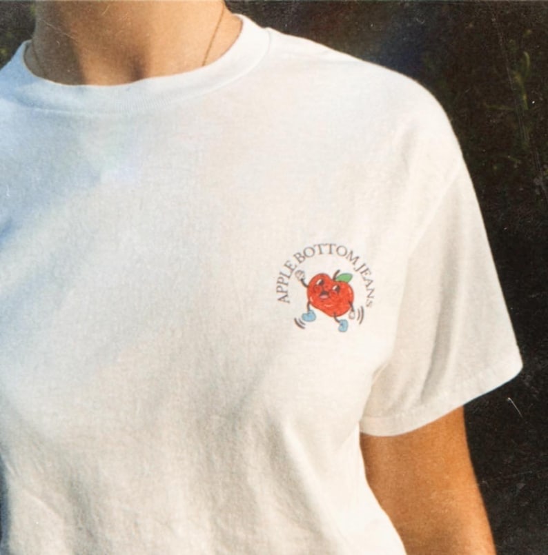Front of white t-shirt with a small apple graphic on the chest and the text “Apple Bottom Jeans” in retro font.