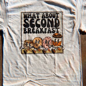 Unisex beige shirt with a retro graphic of breakfast foods and “What About Second Breakfast” text, perfect for LOTR fans.