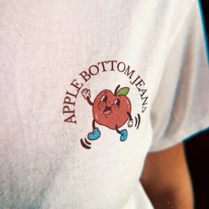 close up of the front graphic on the chest of a retro apple dancing and the text “Apple Bottom Jeans”