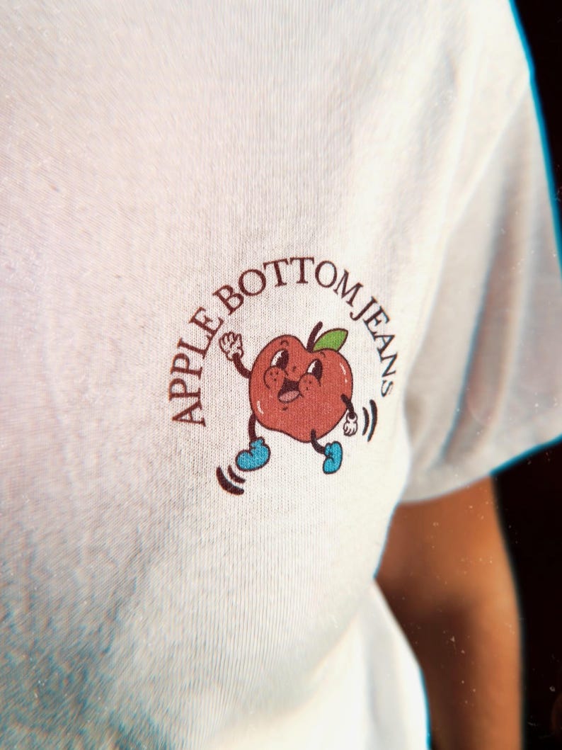 close up of the front graphic on the chest of a retro apple dancing and the text “Apple Bottom Jeans”