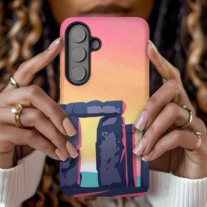 May include: A pink, orange and blue phone case featuring a graphic of Stonehenge at sunset. The case has a cutout for a camera lens with three holes.