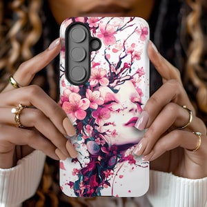 May include: A white phone case featuring a floral design with pink cherry blossoms and a woman's face. The case has a triple-lens camera cutout and is held by hands with gold rings.