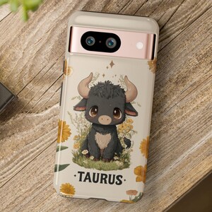 May include: A phone case featuring a cartoon bull illustration with the word "TAURUS." The case has a cream background with yellow flowers and a black bull with large eyes and horns. The phone case is on a wooden surface.