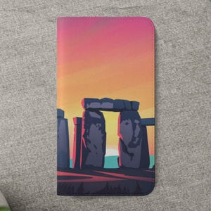 May include: A passport holder featuring a stylized illustration of Stonehenge at sunset. The design includes the iconic stone circle in shades of gray and blue against a vibrant orange and pink sky. The holder is rectangular and made of a smooth material.