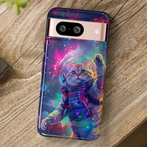 May include: A phone case featuring a cat in an astronaut suit against a vibrant nebula background. The case has a dark blue border and a gold-colored camera housing. The cat is wearing a space helmet and waving.