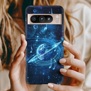 Op de afbeelding: Donkerblauwe telefoonhoes met een hemels ontwerp met een planeet omcirkeld door een gloeiende ring en muzieknoten. De hoes heeft een goudkleurige camerabalk en is ontworpen voor een smartphone.