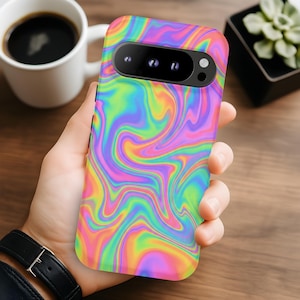May include: A smartphone case with a vibrant, swirling pattern in shades of pink, yellow, green, and blue. The case has a camera cutout and is held in a person's hand. The background includes a coffee cup and a small plant.