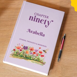 Könnte beinhalten: Ein hellviolettes Notizbuch mit dem aufgedruckten Text Chapter ninety, Arabella und Stories, Thoughts & Memories auf dem Einband. Der Einband zeigt unten eine farbenfrohe Aquarellillustration verschiedener Blumen und einer Biene, dazu ein Stift.