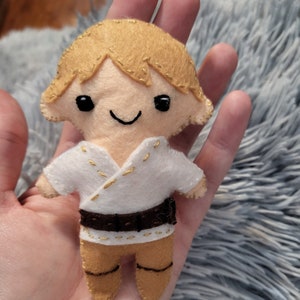 May include: A small felt doll with blonde hair, wearing a white shirt with gold stitching, a brown belt, and brown pants. The doll has a smiling face with black eyes.