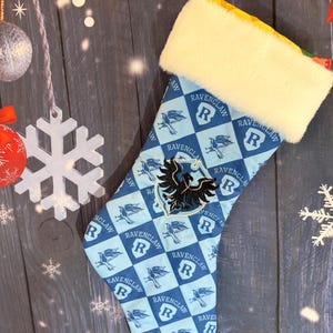 Peut inclure: Une chaussette de Noël avec un motif à carreaux bleu clair et bleu foncé, ornée de blasons Ravenclaw et d'illustrations d'oiseaux. Elle présente un revers en fausse fourrure blanche et un emblème d'aigle noir brodé en son centre.