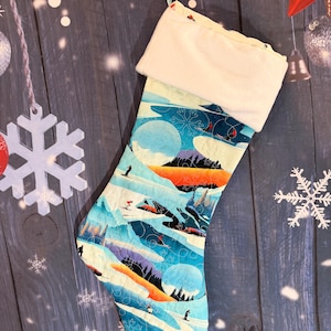 Op de afbeelding: Een kerstkous met een kleurrijke berglandschap-print met skiërs, besneeuwde bergtoppen en dennenbomen in tinten blauw, oranje en wit. De kous heeft een effen crèmekleurige pluche boord aan de bovenkant en een klein lusje om op te hangen.