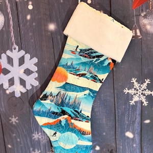 Op de afbeelding: Een kerstkous met een gewatteerd stoffen patroon van een winters berglandschap met skiërs, dennenbomen en grote cirkelvormige zonnen in tinten blauwgroen, oranje en wit. De kous heeft een pluche, effen witte boord en een stoffen lus.