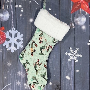 May include: A Christmas stocking featuring a light green fabric with a repeating pattern of mermaids wearing Santa hats and holding tridents. The stocking has a thick, white faux fur cuff at the top and a small fabric loop for hanging.